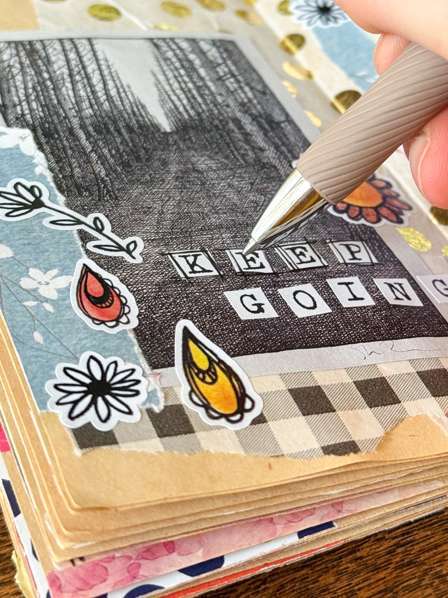 Hand holding a pen over a scrapbook page with typewriter alphabet stickers saying 'KEEP GOING' text and decorative elements in a junk journal.