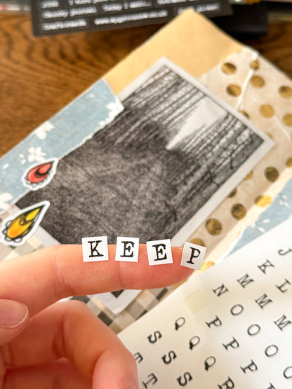 Hand holding small typewriter alphabet stickers spelling 'KEEP' over a collage of images and text in a junk journal.