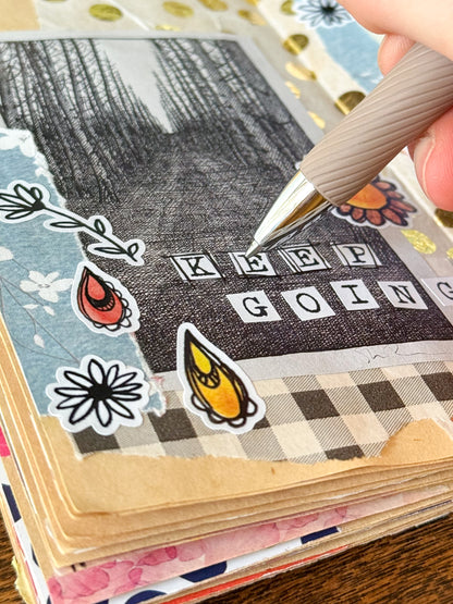 Hand holding a pen over a scrapbook page with typewriter alphabet stickers saying 'KEEP GOING' text and decorative elements in a junk journal.